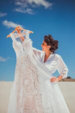 bride-to-be holding a beautiful handmade lace wedding dress against a sunny blue sky and a long sandy beach