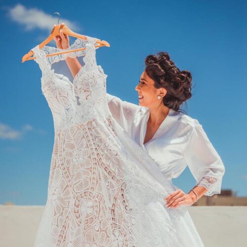bride-to-be holding a beautiful handmade lace wedding dress against a sunny blue sky and a long sandy beach